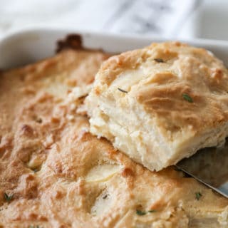 A white pan of potatoes au gratin with a square of the potatoes on a spatula being removed from the pan.