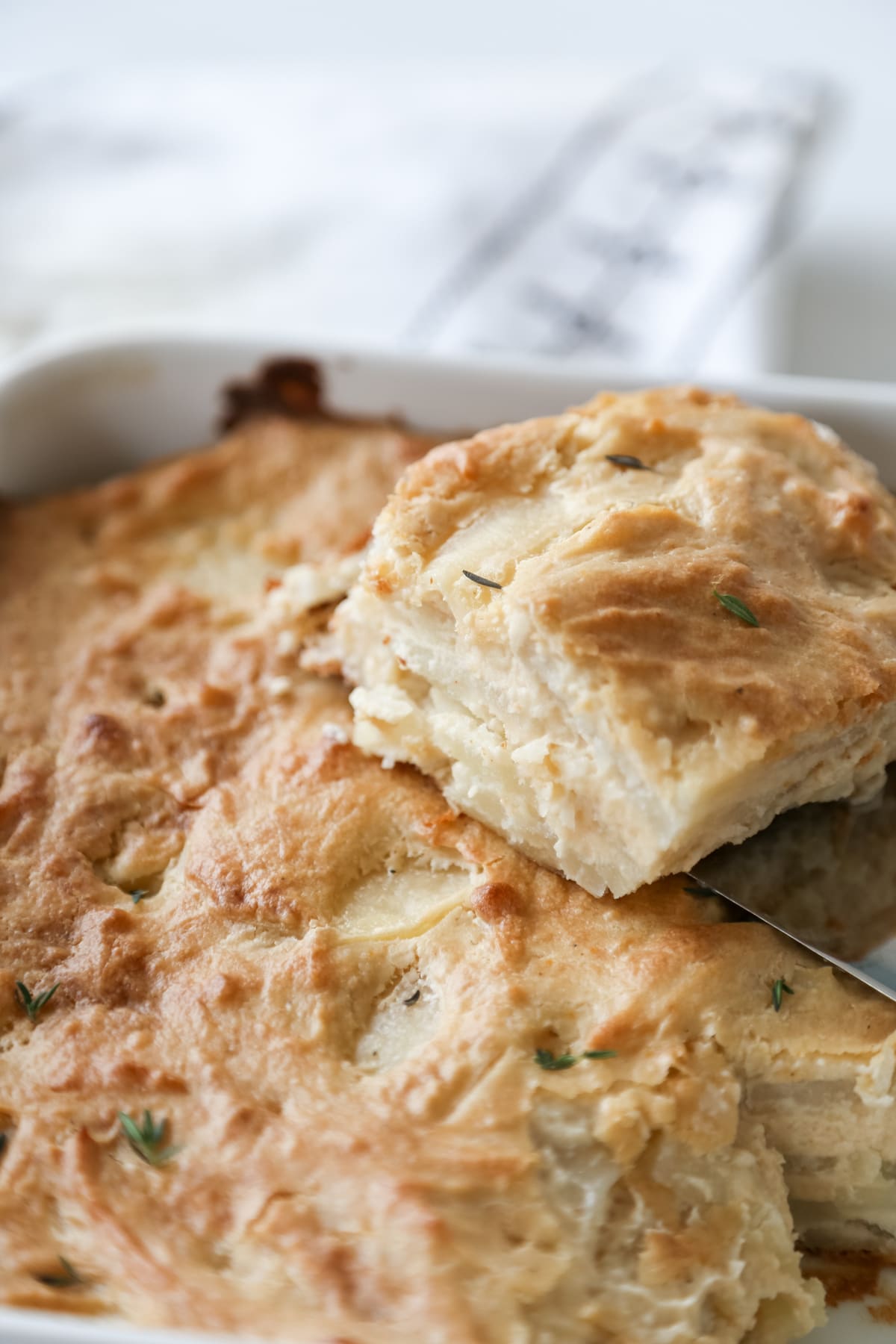 A white pan of potatoes au gratin with a square of the potatoes on a spatula being removed from the pan.