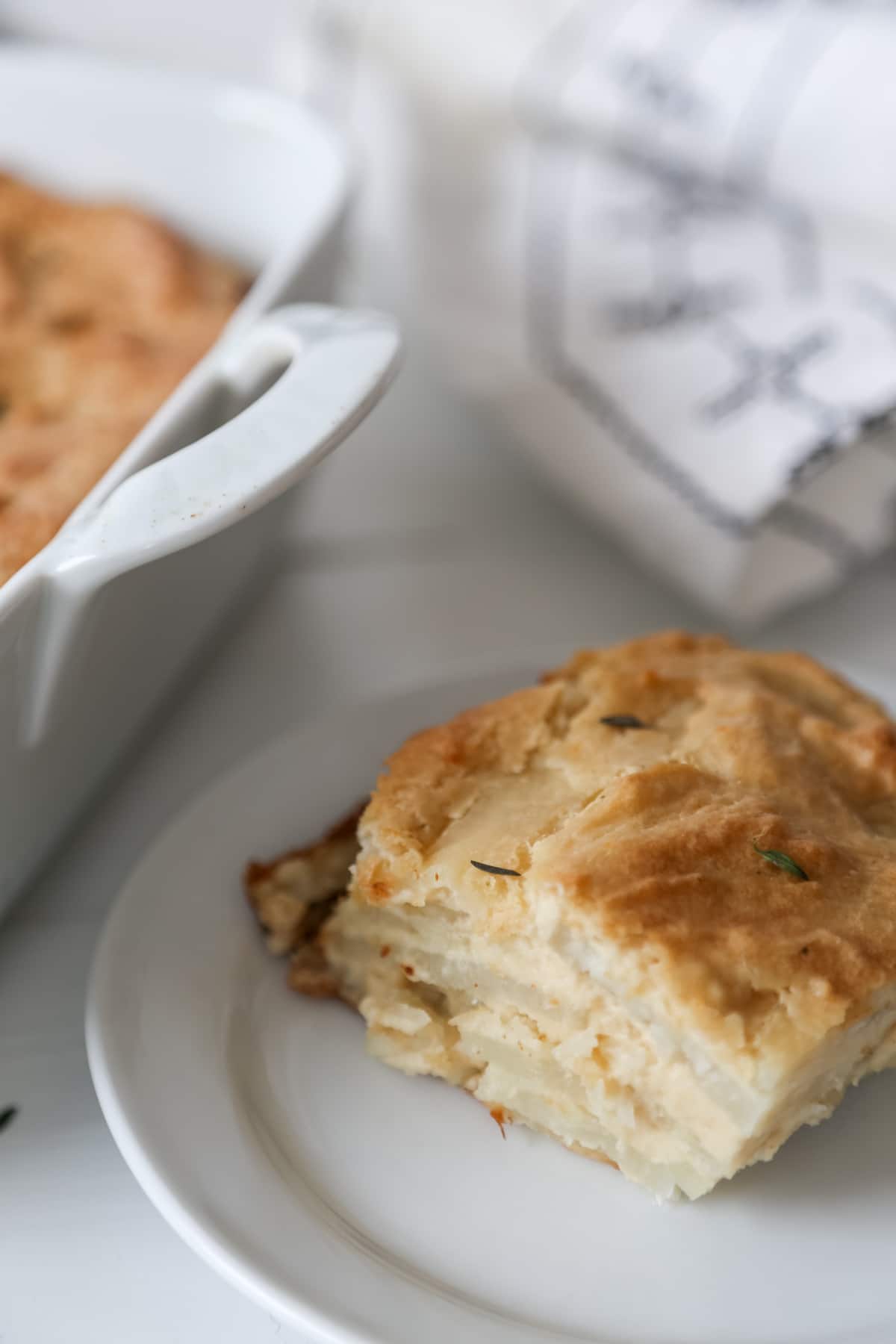 A white plate with a square of potatoes au gratin and the white dish of the potatoes next to it.