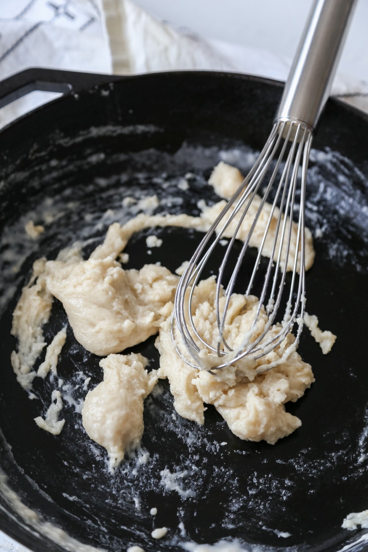 A cast iron skillet with a silver whisk that's whisking a flour and oil roux into a thick paste.