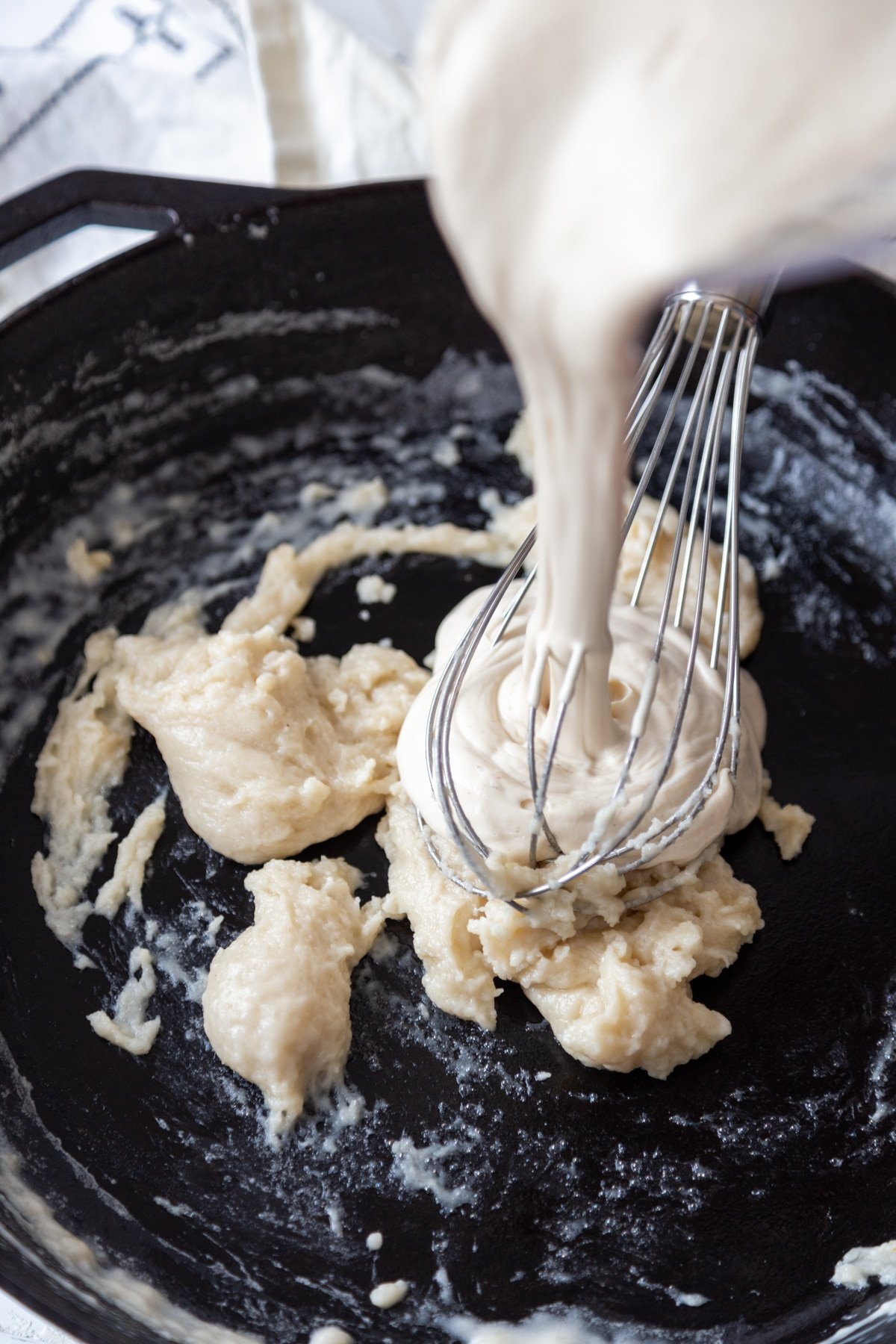 A cast iron skillet with a roux and a white cream sauce being whisked into the pan.