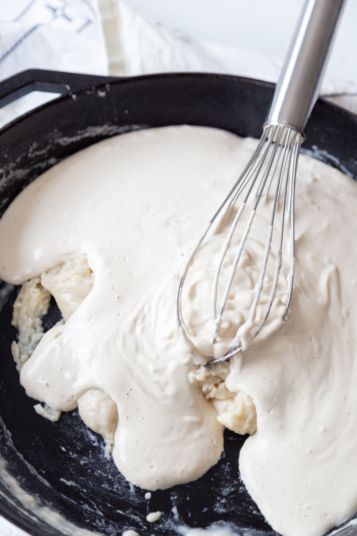 A silver whisk is whisking a roux and cream sauce together.