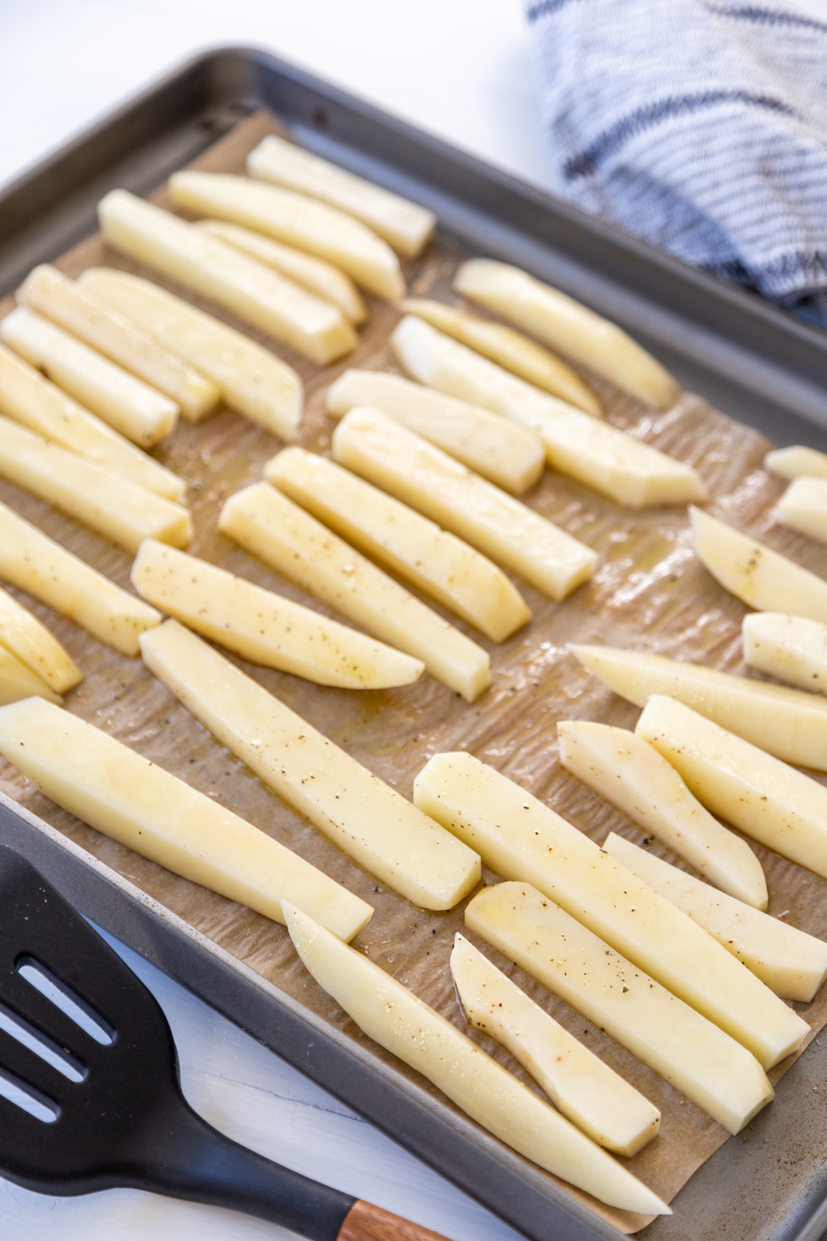 A rimmed baking sheet with unbaked French fries on brown parchment paper and a black spatula and a white and black towel next to the baking sheet.