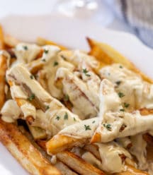 A white oval platter with fries that are covered in cheese sauce and gravy, and sprinkled with thyme leaves.