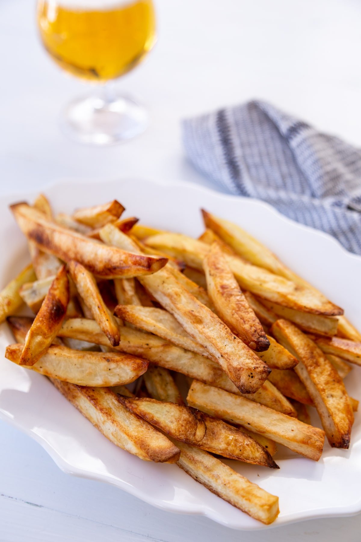 A white oval scalloped edge platter with a pile of fries and a glass of beer and a black and white towel next to the platter.