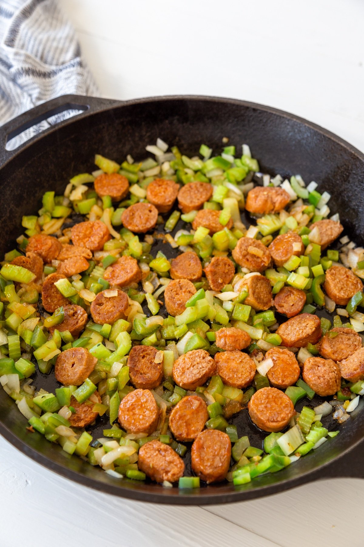 Cooked sliced sausage, onion, green bell pepper, and celery in a black iron skillet with a white and black towel next to the pan.