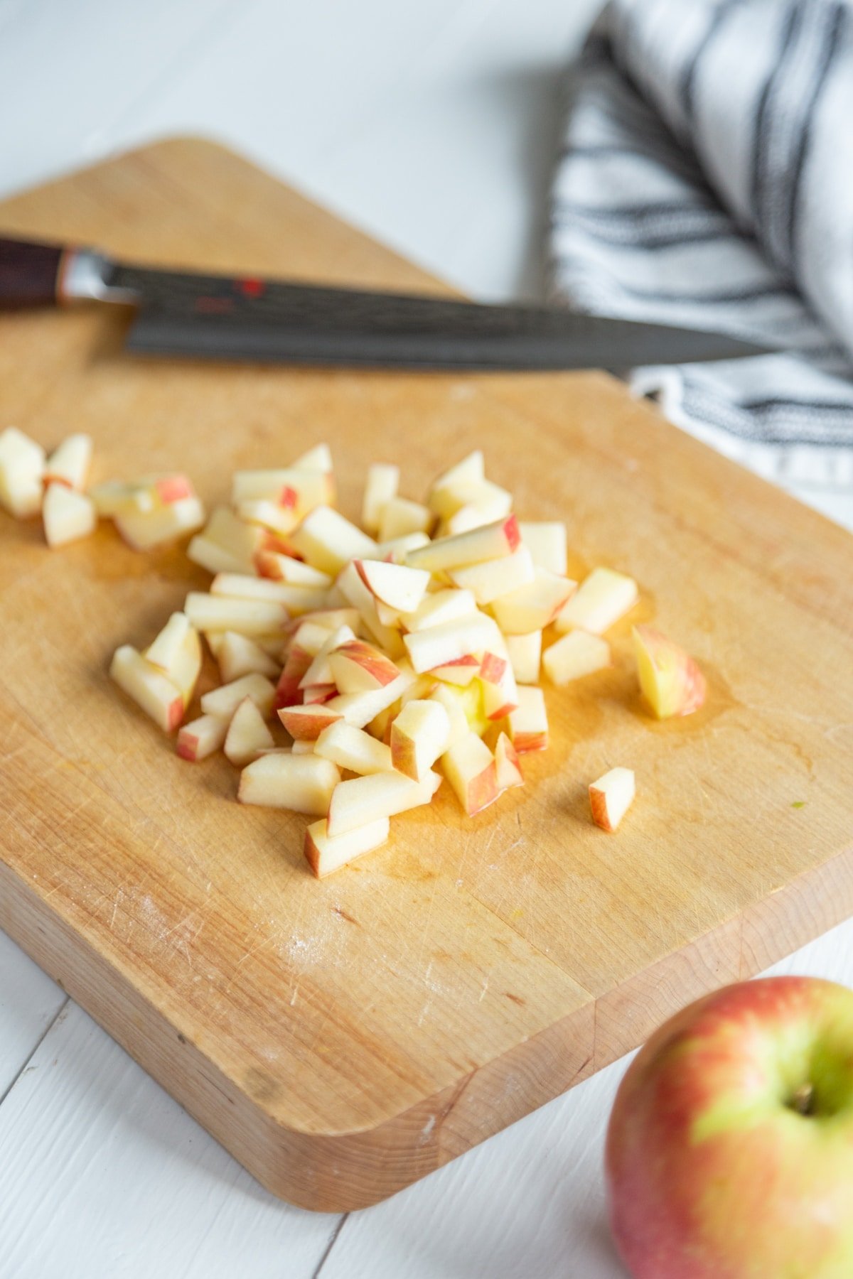 A wood cutting board with a knife and chopped apples and a whole red apple and a towel next to the board.