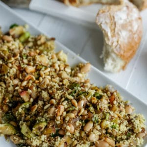 A rectangular white platter with a couscous and Brussels sprouts salad and a loaf of bread with a piece torn off of it next to the platter.