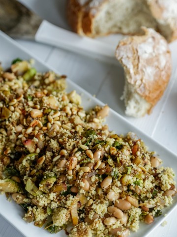 A rectangular white platter with a couscous and Brussels sprouts salad and a loaf of bread with a piece torn off of it next to the platter.