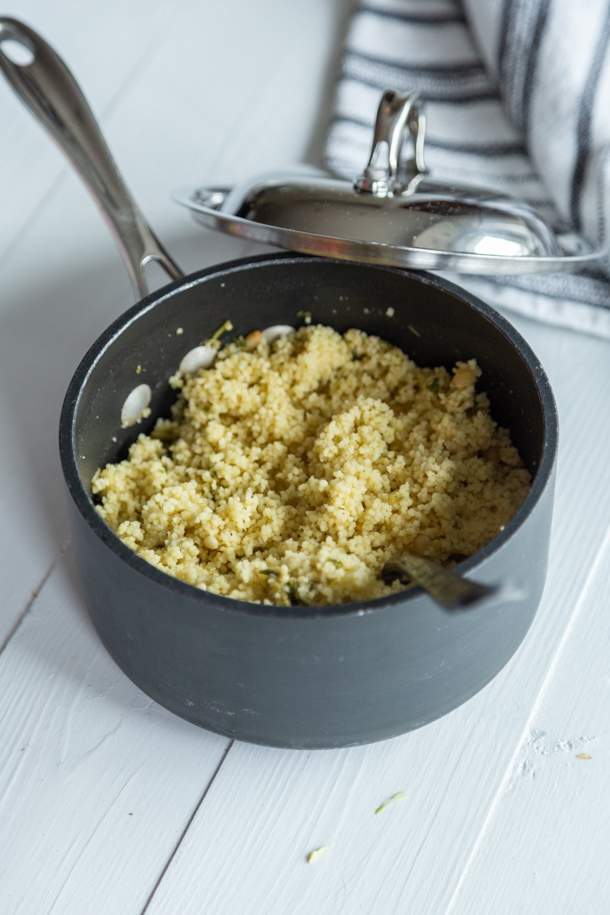 A saucepan with cooked couscous on a white wood table with a white and black striped towel next to the pan. 