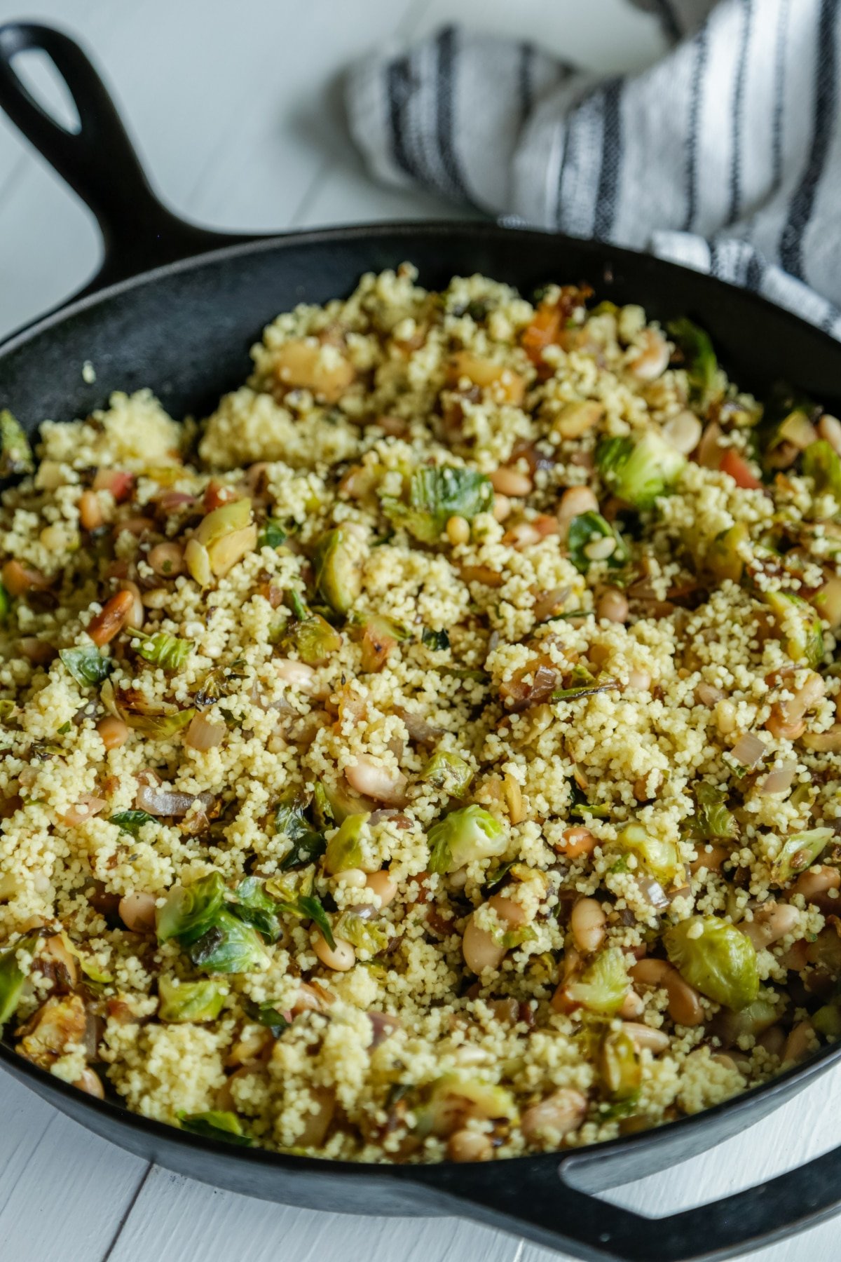 An iron skillet with warm couscous and Brussels sprouts salad and a white and black striped towel next to the pan.