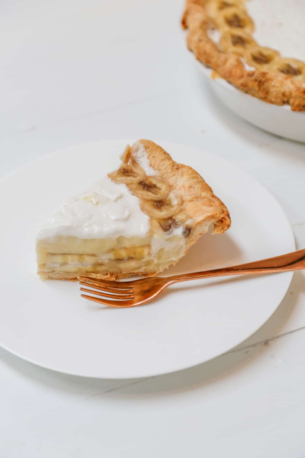 A slice of banana cream pie on a white plate with a copper fork on the plate, and the whole pie in the background.