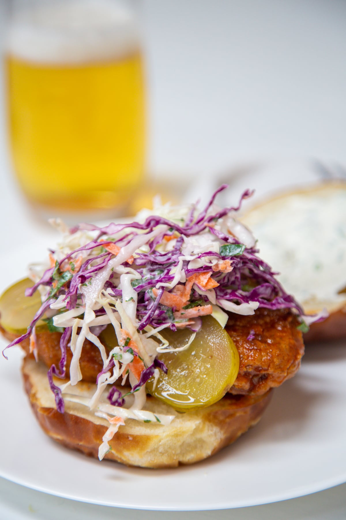 A fried chicken sandwich with pickles and creamy coleslaw on top of it and a glass of beer in the background.