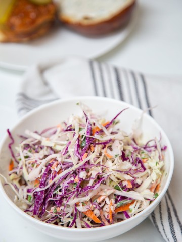 Red and green cabbage creamy coleslaw in a white bowl next to a white and black striped towel with a bun in the background.