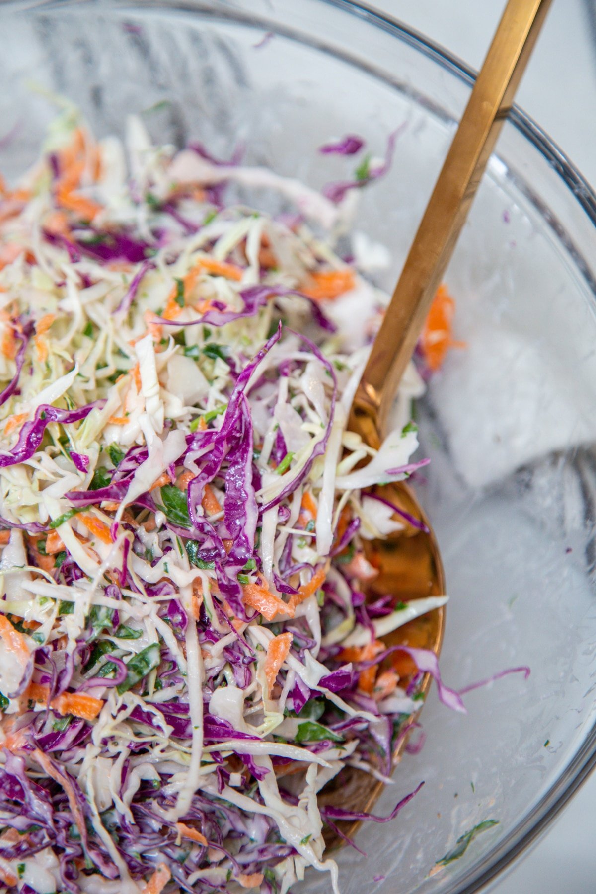 Gold salad serving pieces tossing creamy coleslaw with red and green cabbage in a clear glass bowl. 