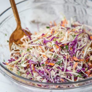 A clear glass bowl with creamy coleslaw with purple and green cabbage and carrots, and gold serving pieces in the bowl.