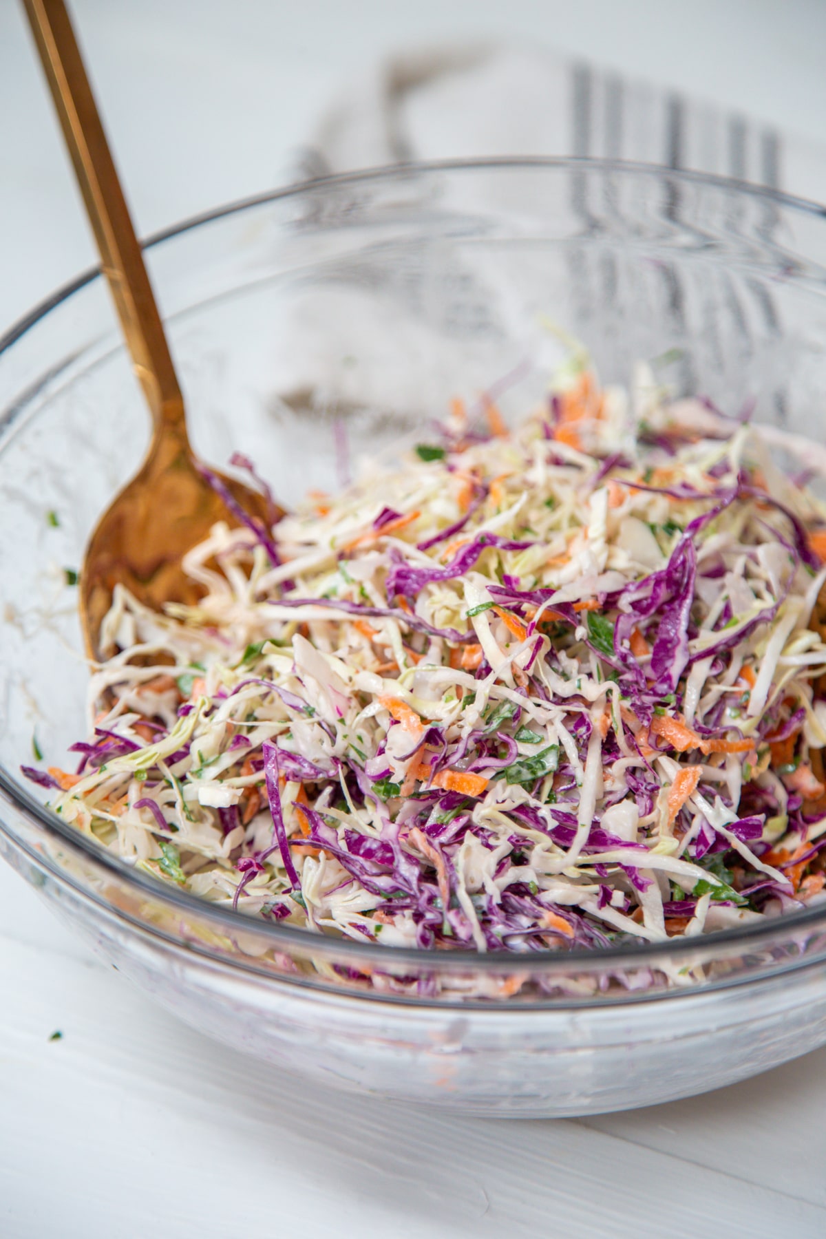 A clear glass bowl with creamy coleslaw with purple and green cabbage and carrots, and gold serving pieces in the bowl.