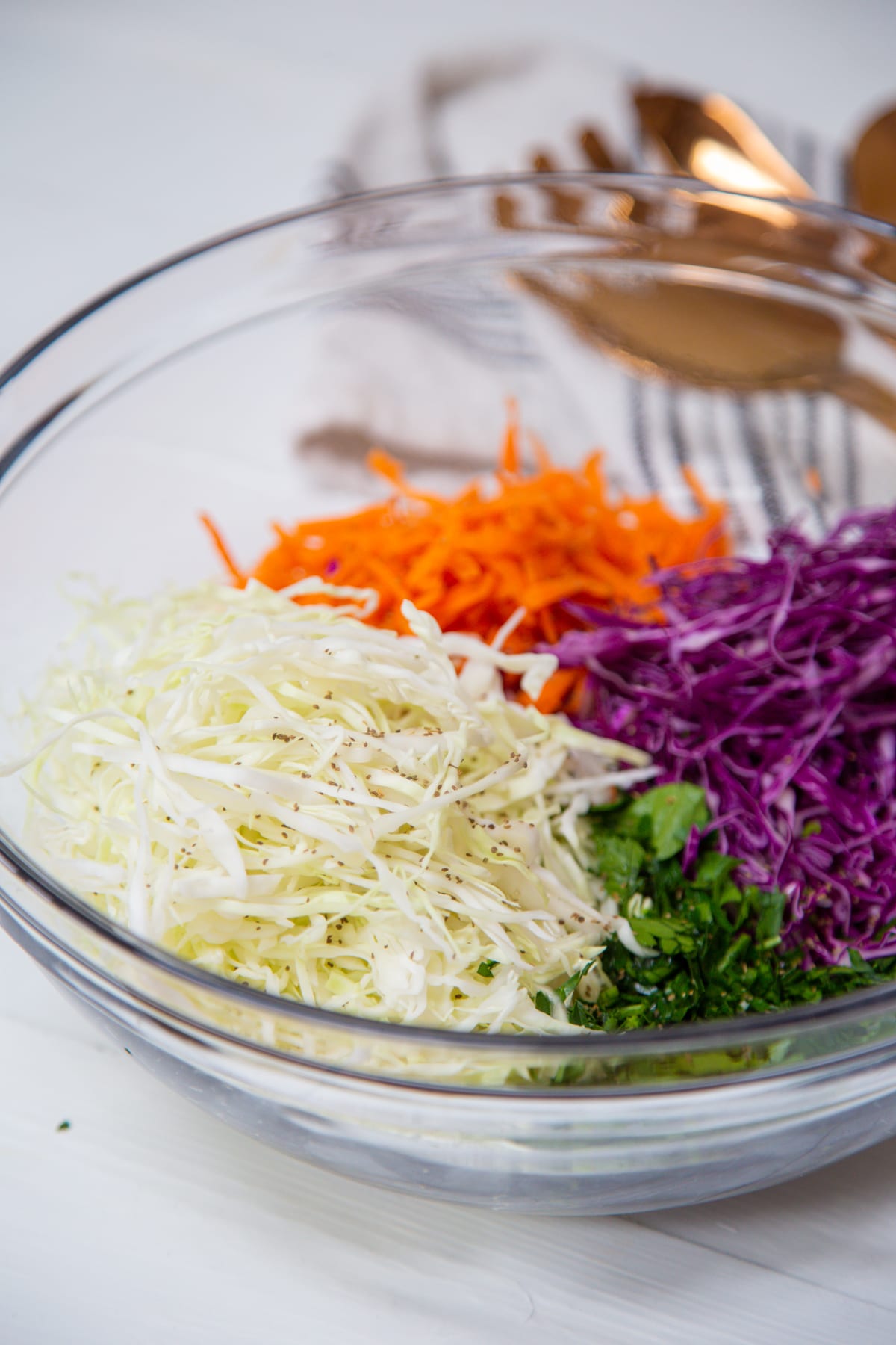 A clear glass bowl filled with shredded green and red cabbage, grated orange carrots, and chopped parsley.