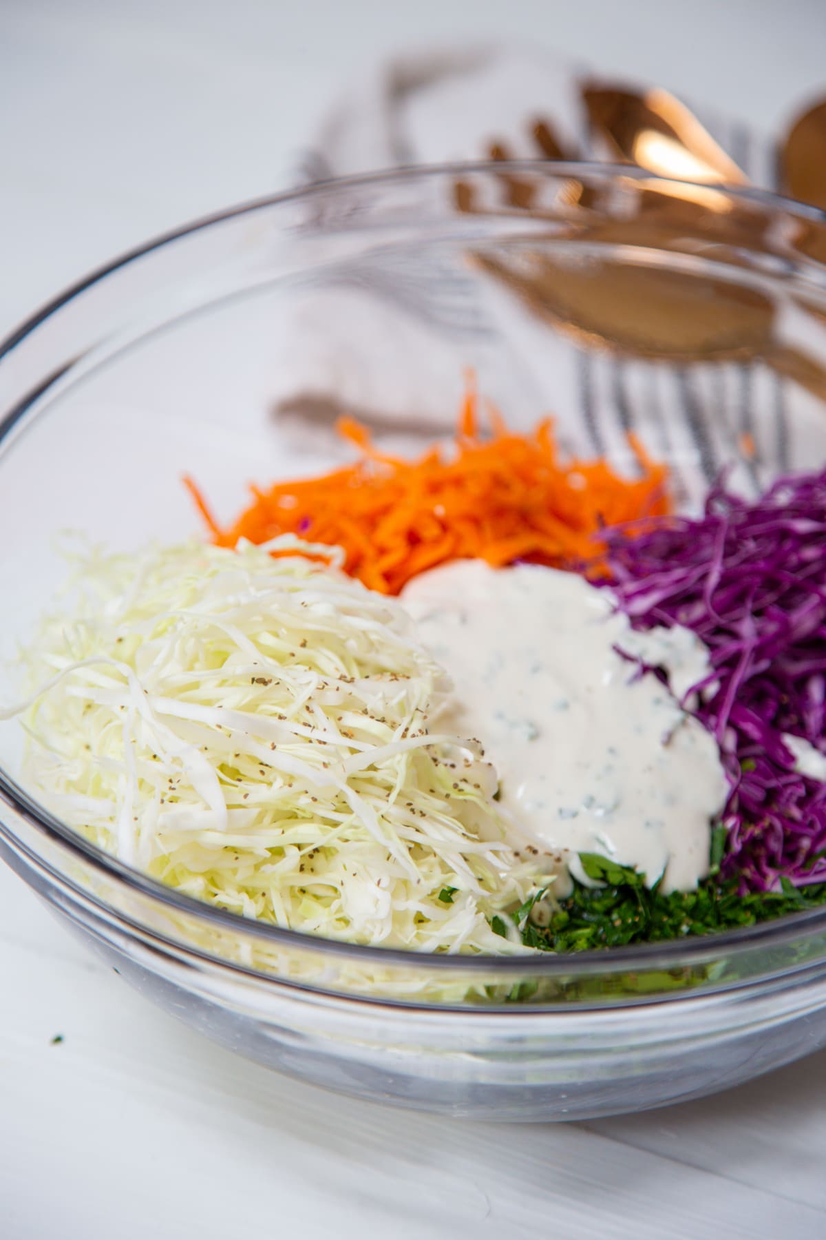 A clear glass bowl with shredded green and purple cabbage, grated carrots, chopped parsley, and a white creamy dressing. 