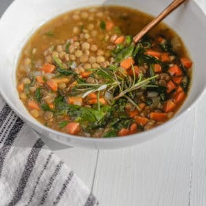 A white bowl with vegetable lentil soup with a copper spoon in the bowl and a white and black towel next to the bowl.