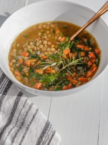 A white bowl with vegetable lentil soup with a copper spoon in the bowl and a white and black towel next to the bowl.