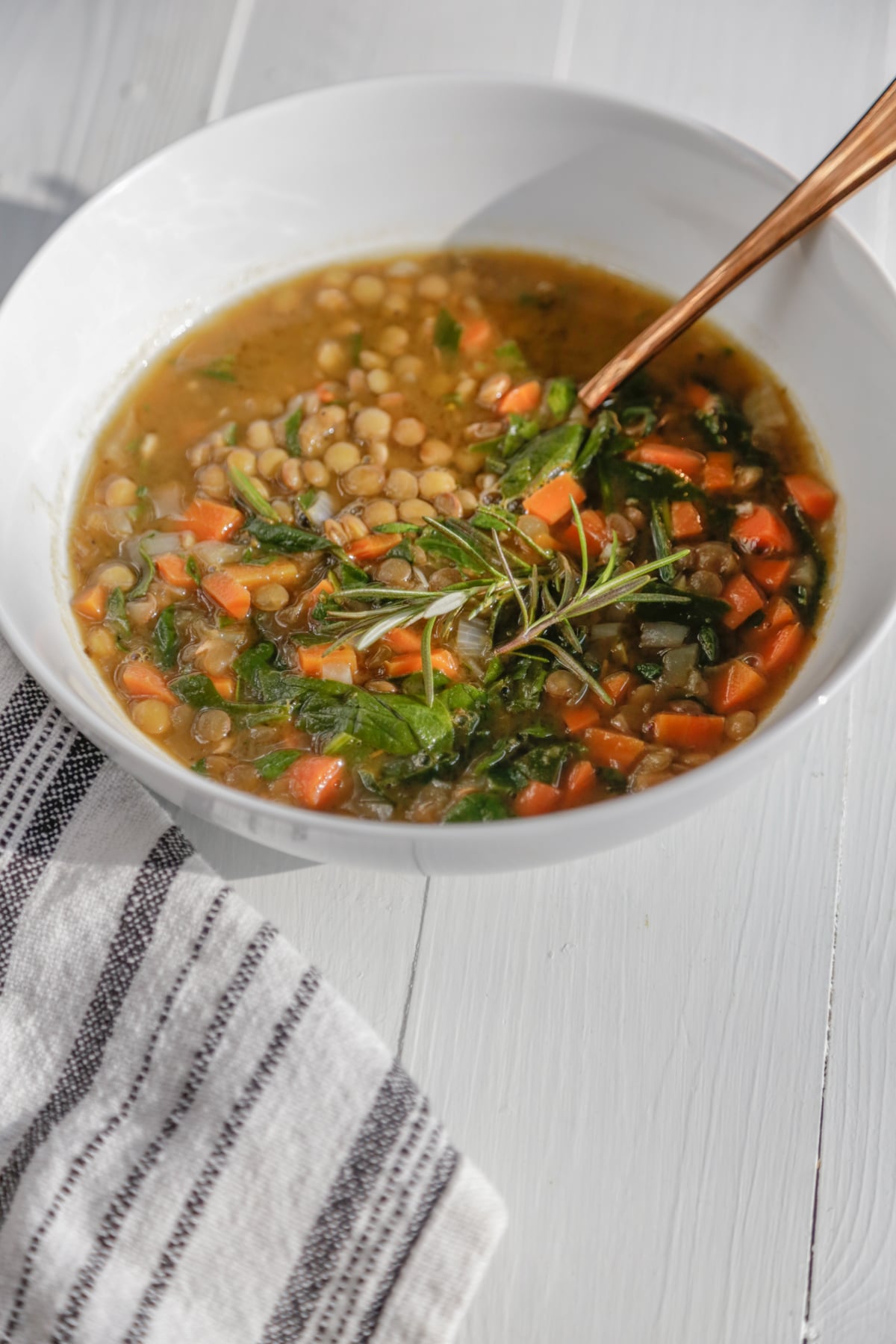 A white bowl with vegetable lentil soup with a copper spoon in the bowl and a white and black towel next to the bowl.