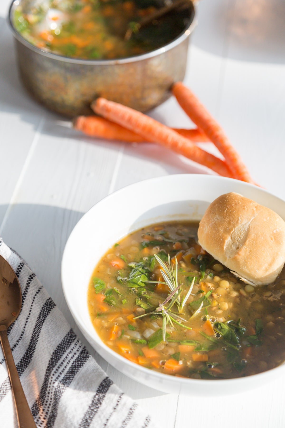 A white bowl with lentil and vegetable soup with a roll of bread in the bowl and a stack of carrots on a white table next to a copper pot of the soup.