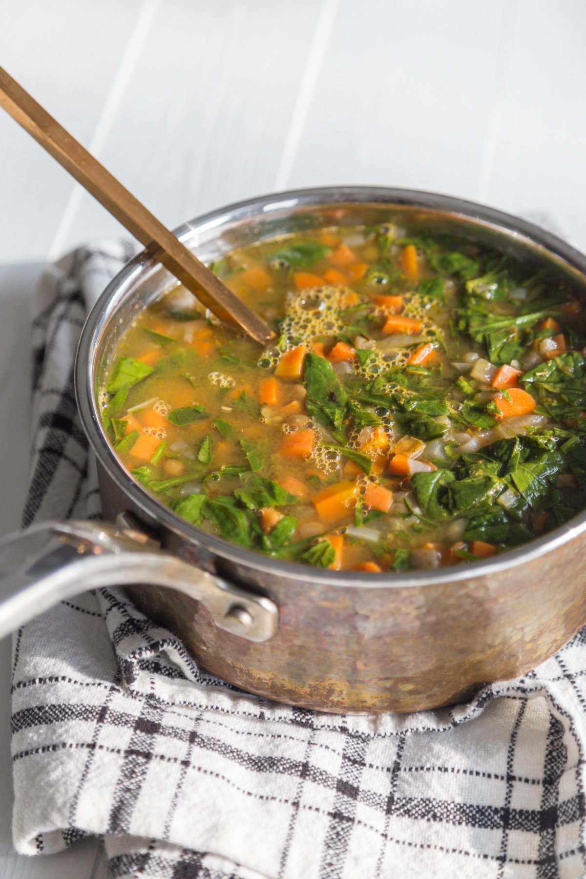 Vegetable soup in a copper pot that's on a black and white towel.