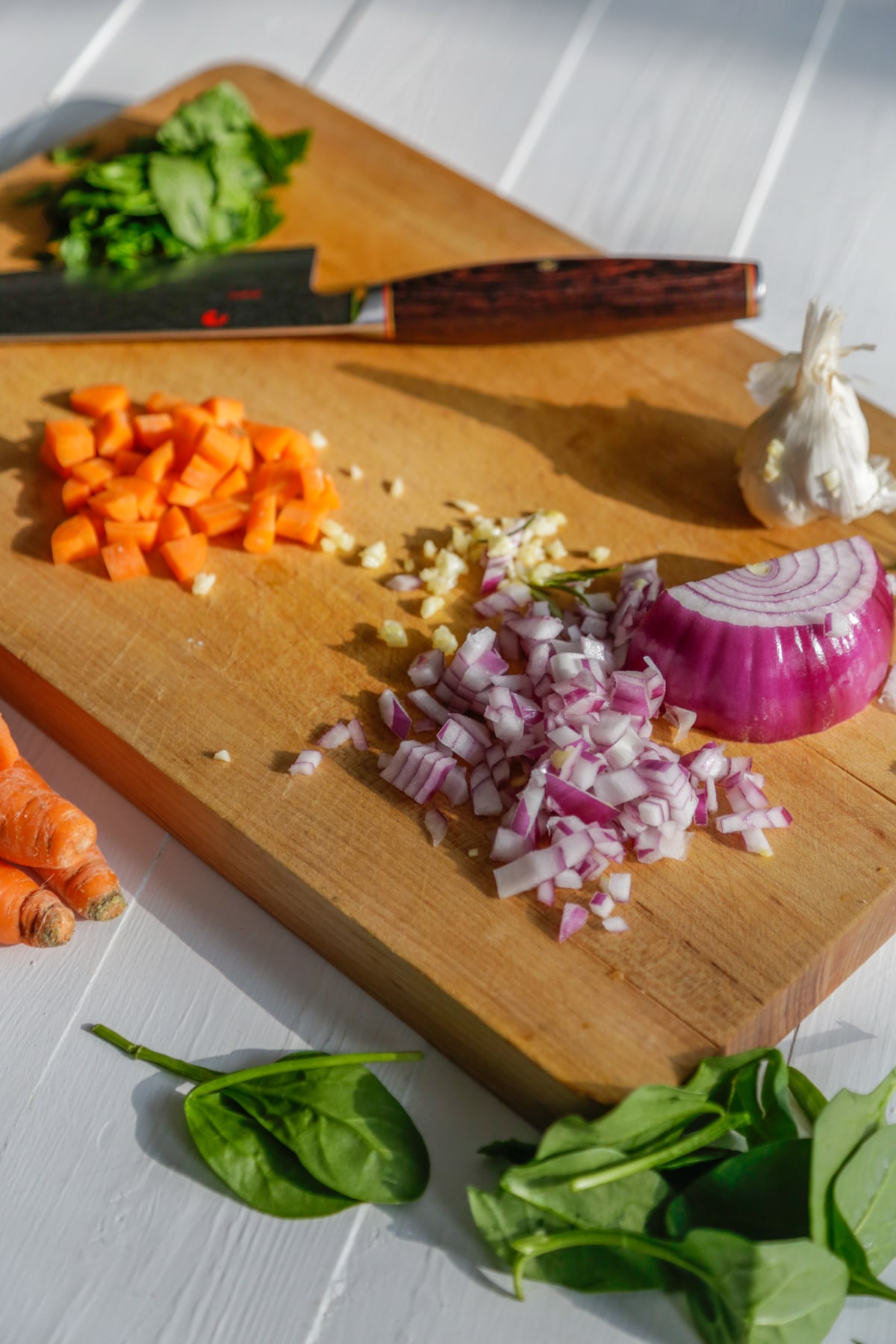 A wood cutting board with diced red onion, carrots, garlic and spinach.