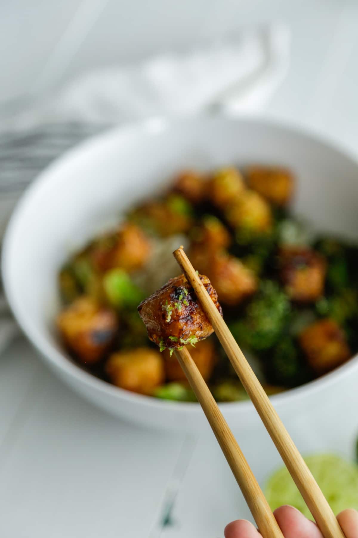 A pair of chopsticks holding a piece of tofu over a white bowl filled with the tofu, broccoli, and rice.