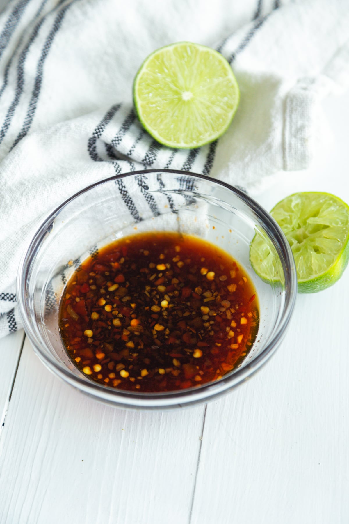 A glass bowl with soy garlic sauce and a white and black striped towel and lime halves next to the bowl.