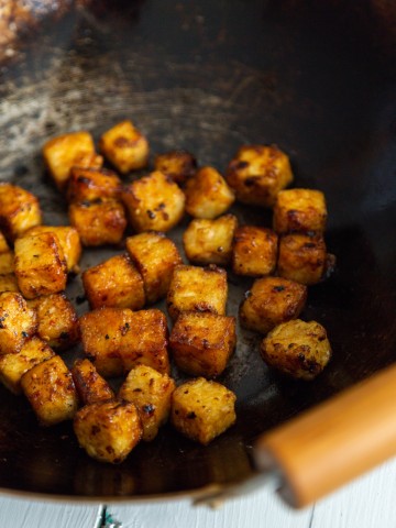A close up picture of cubed fried tofu in a wok with wooden handles.