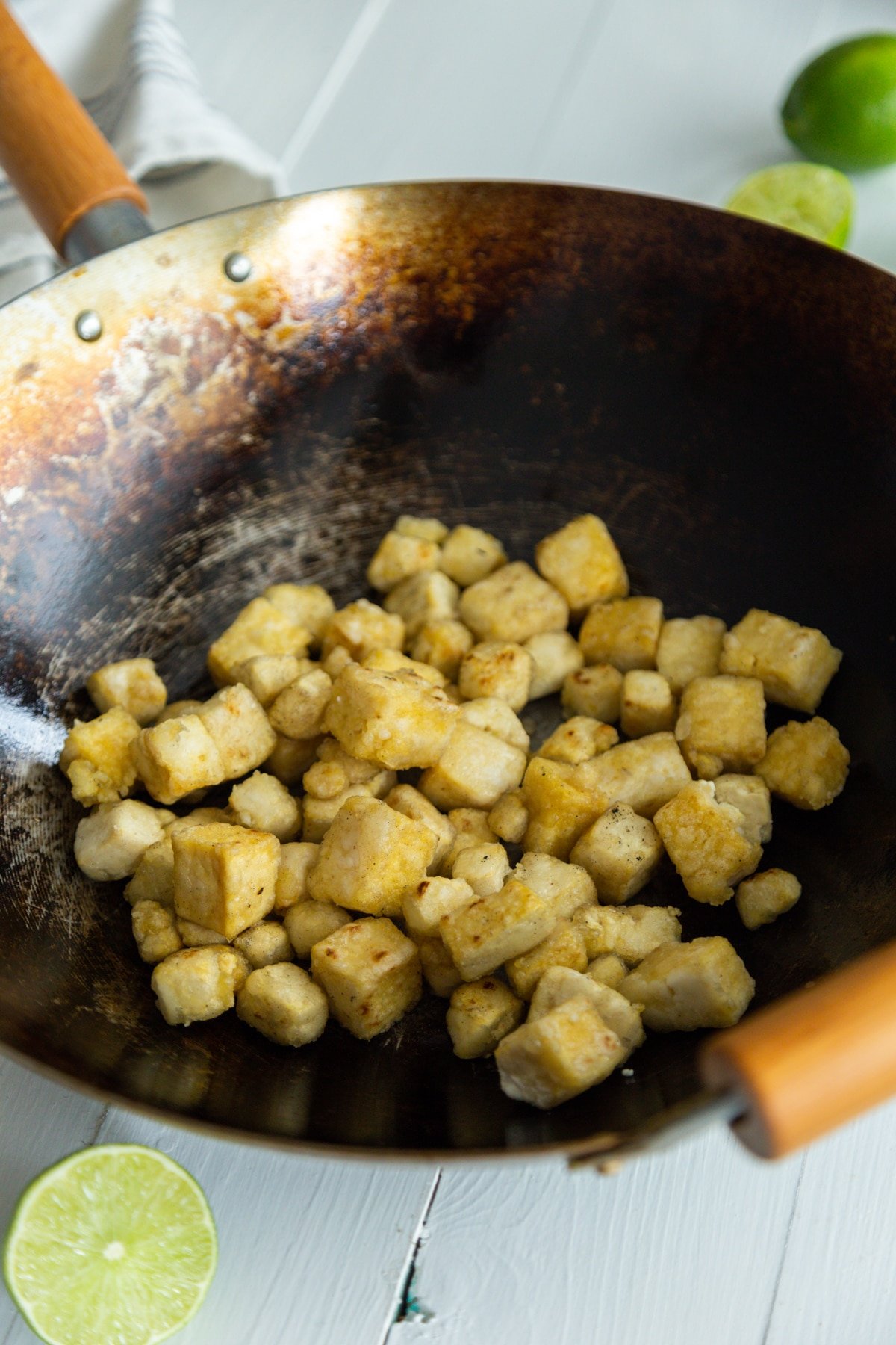 A wok with cubed crispy tofu on a white wood table with limes next to the pan.