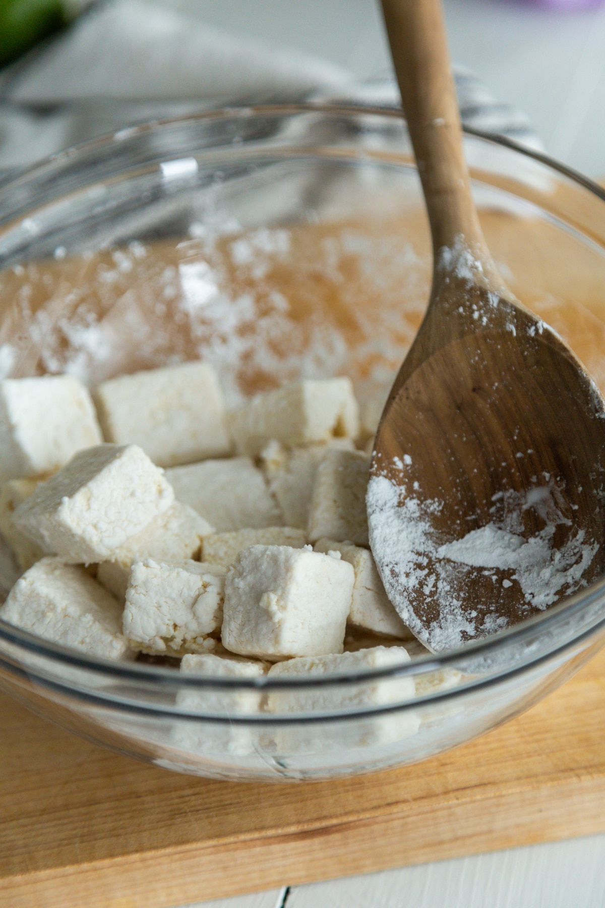 A glass bowl with cubed tofu and corn starch being tossed with a wooden spoon.