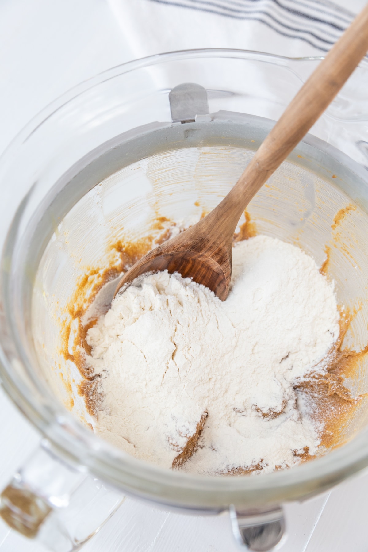 A glass mixing bowl with whipped butter and sugar and a wooden spoon stirring in the flour.