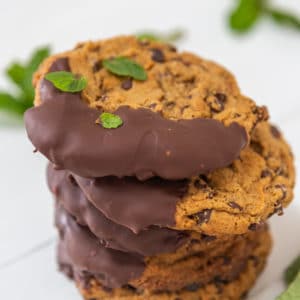Four chocolate dipped chocolate chip cookies stacked with mint leaves next to them.