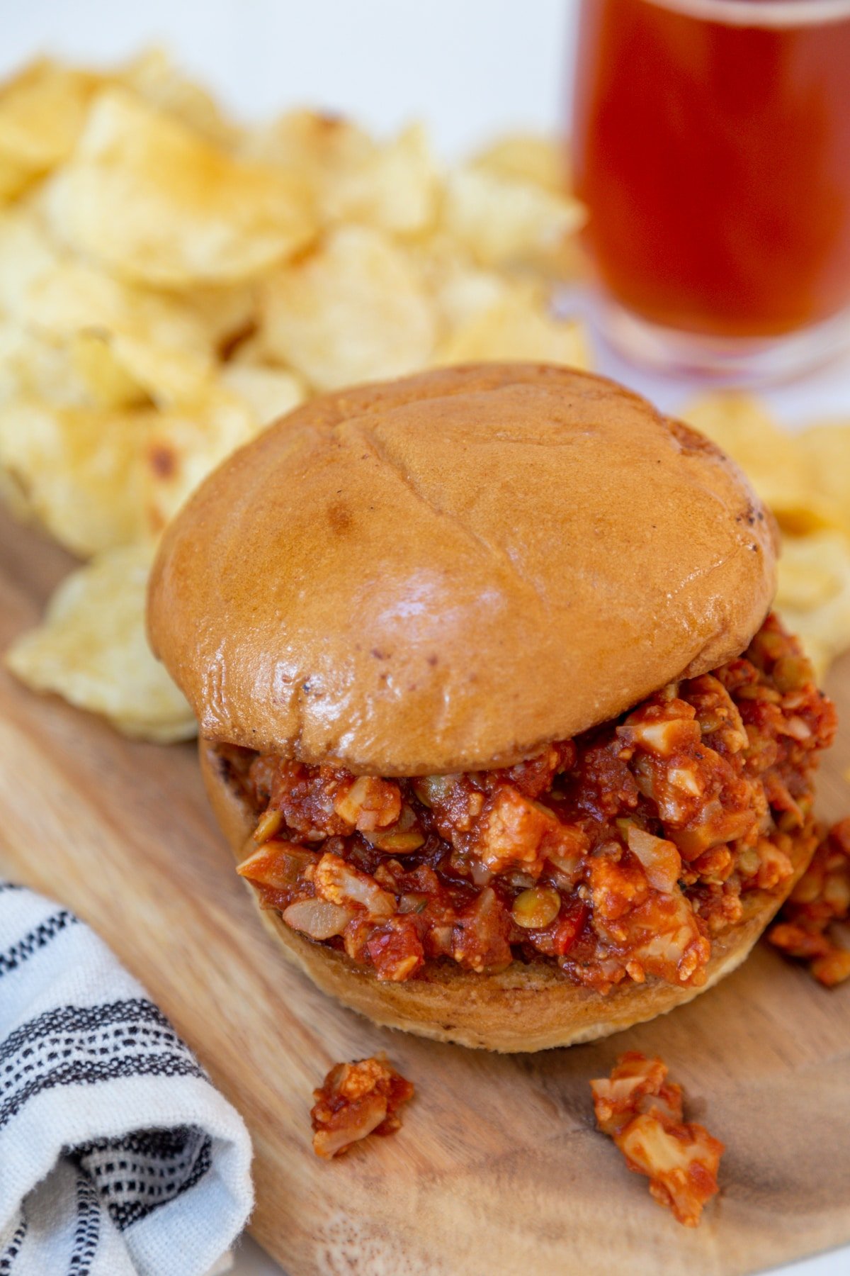 A sloppy Joe sandwich on a wooden board with potato chips and a glass of beer in the background.