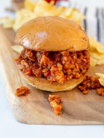 A wooden board with a sloppy Joe sandwich and potato chips.