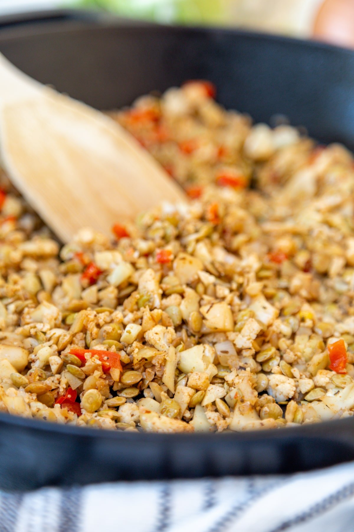 An iron skillet with lentils and finely chopped cauliflower, onion, and bell pepper being stirred with a wooden spoon.