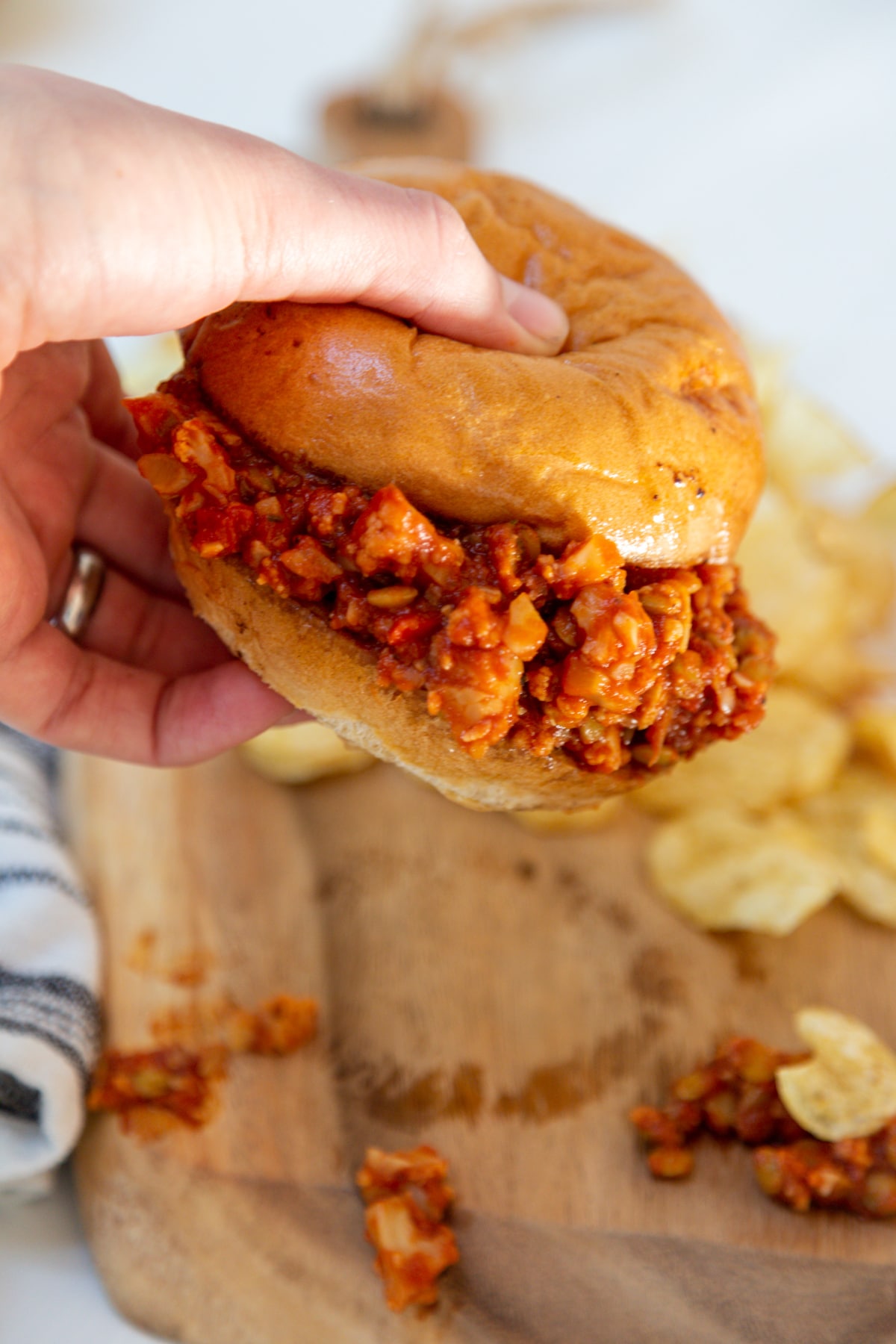 A hand holding up a sloppy Joe sandwich over a wooden board with potato chips on it.