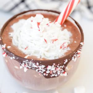 A clear glass mug rimmed with chocolate and crushed peppermint and filled with hot chocolate and whipped cream, and a peppermint stick in the mug.