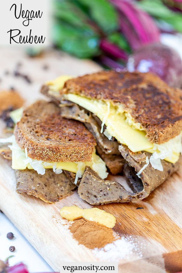 A Pinterest pin for a vegan Reuben with a picture of the sandwich cut in half on a wooden board with spices and a drop of the Russian dressing.