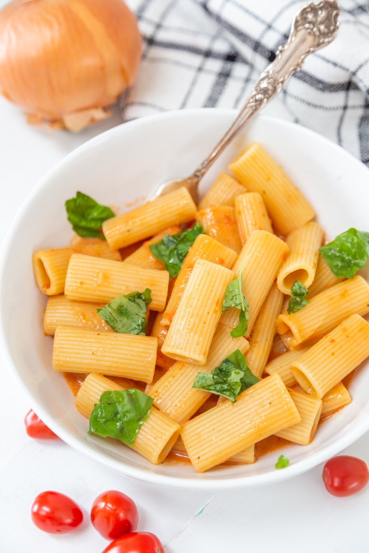 A white bowl with rigatoni and creamy red sauce, with cherry tomatoes and an onion next to the bowl.