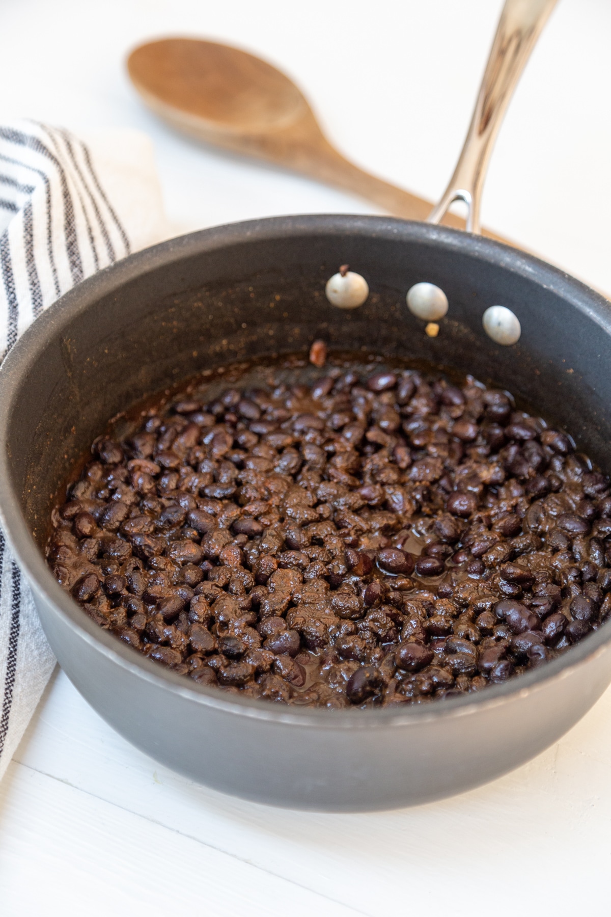A pot of black beans with a wooden spoon and a black and white striped towel in the background.