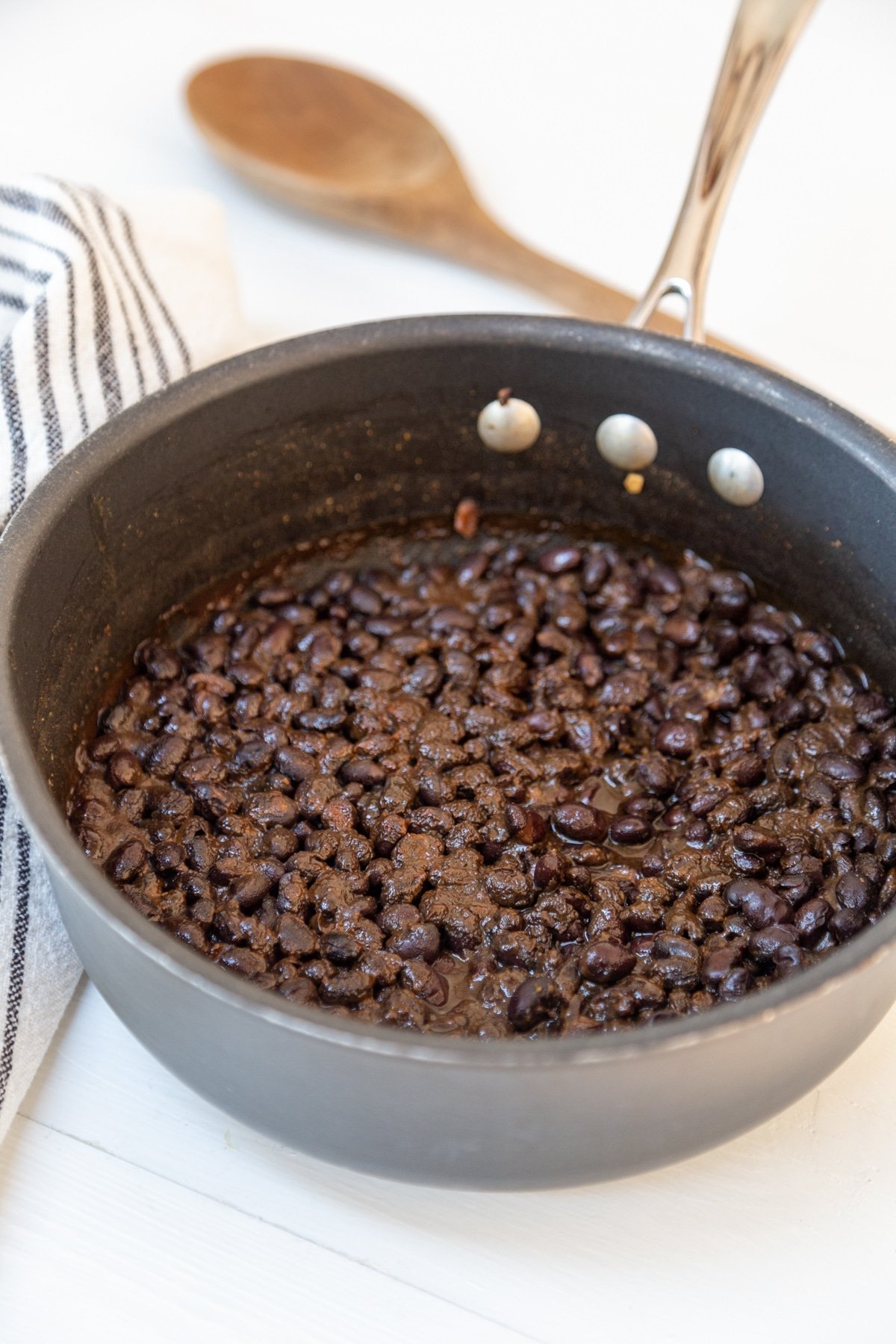 A pot of black beans with a wooden spoon and a black and white striped towel in the background.