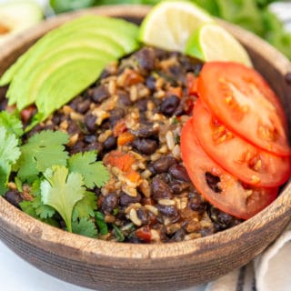 A wooden bowl with black beans, rice, and vegetables, topped with sliced avocados, tomato, and fresh cilantro.