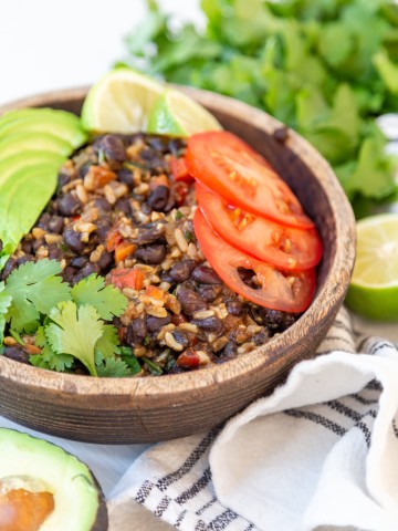 A brown wood bowl with black beans and rice with vegetables and an avocado and cilantro around the bowl.