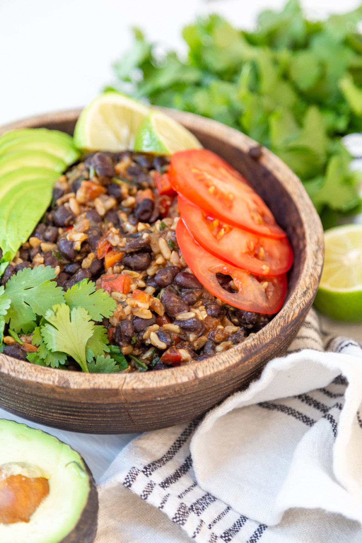 A brown wood bowl with black beans and rice with vegetables and an avocado and cilantro around the bowl. 