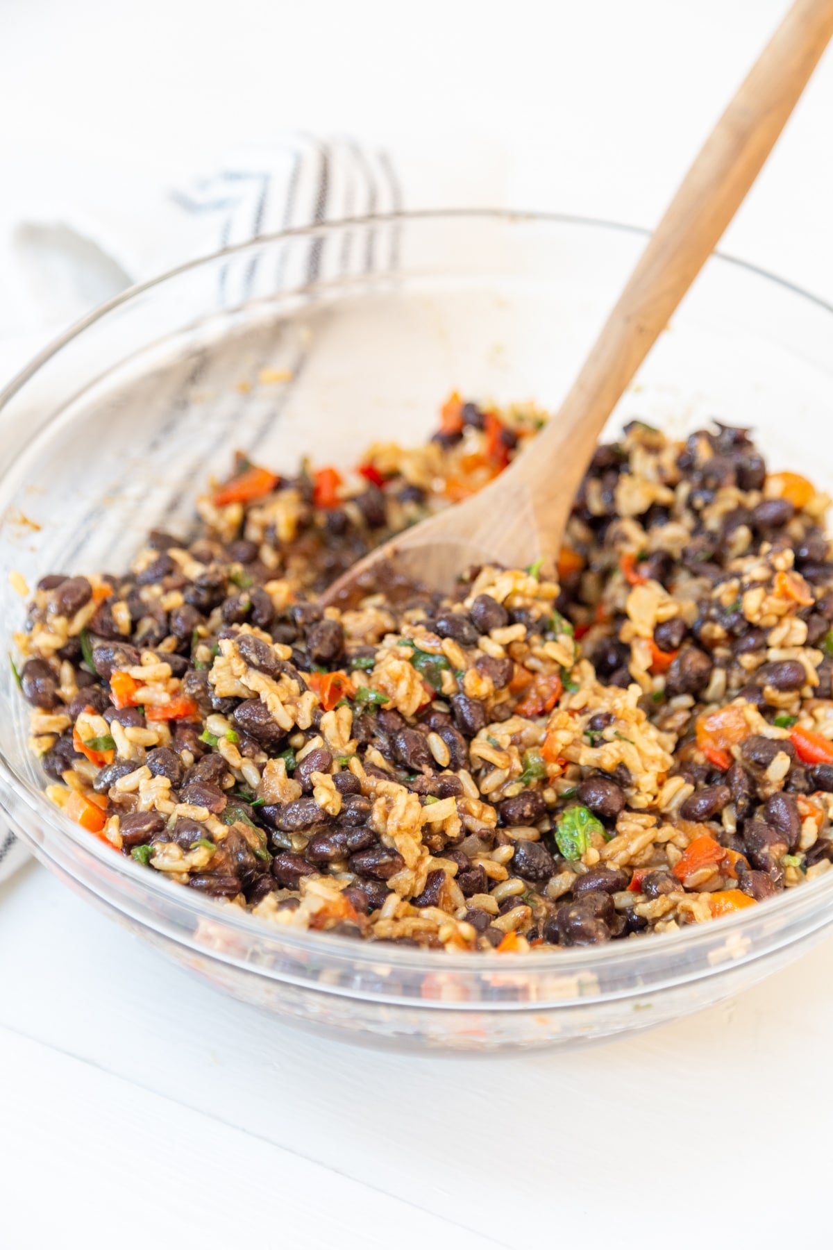 A clear glass bowl with a wooden spoon stirring black beans, rice and mixed vegetables.