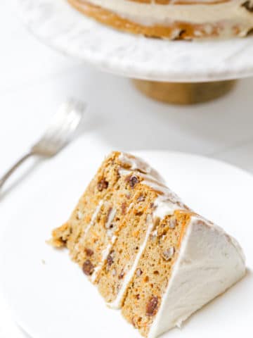A slice of carrot cake with cream cheese frosting on a white plate with the whole cake in the background.